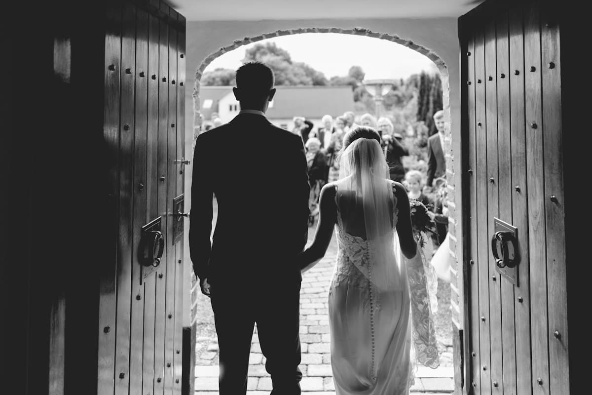 Los novios, de espaldas, salen por una puerta hacia los invitados, en blanco y negro.
